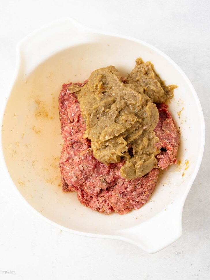A white bowl containing raw ground meat topped with a scoop of brownish-green purée, sitting on a light surface.