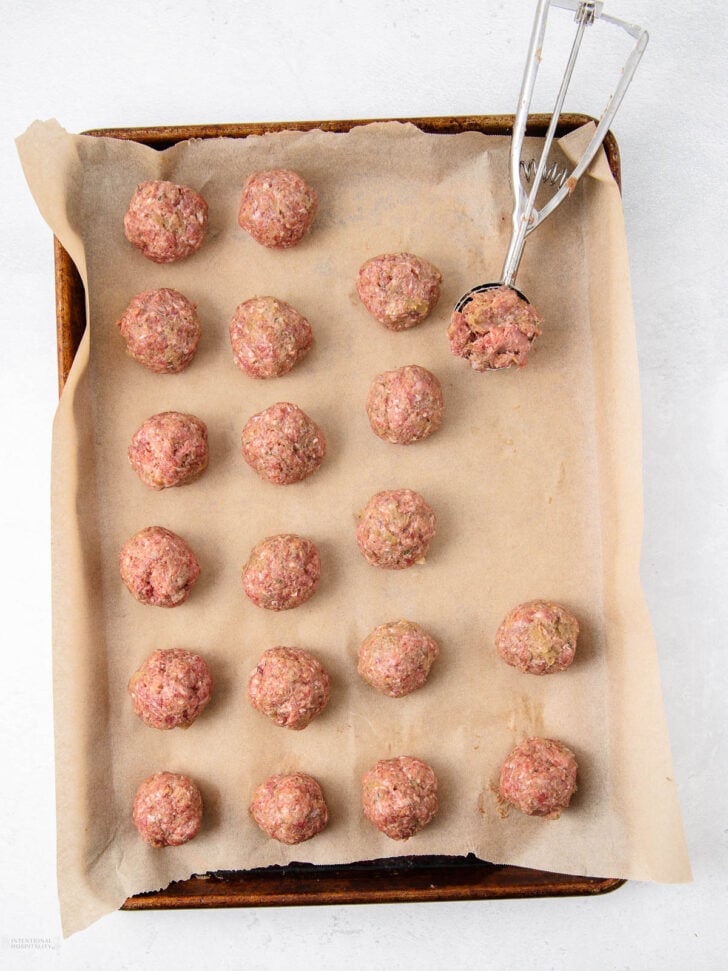 Raw meatballs arranged in rows on a parchment-lined baking sheet. A metal scoop is holding a portion of meatball mixture, ready to form another meatball.