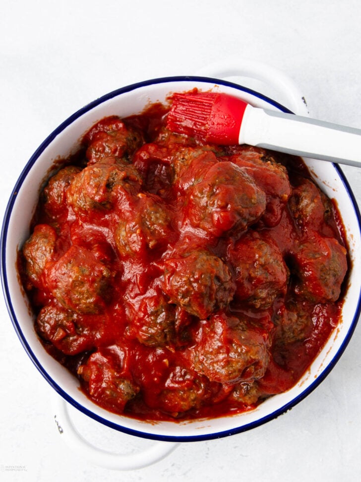 A white enamel bowl filled with meatballs covered in red tomato sauce, with a red and white silicone basting brush resting on the edge. The background is a light, textured surface.