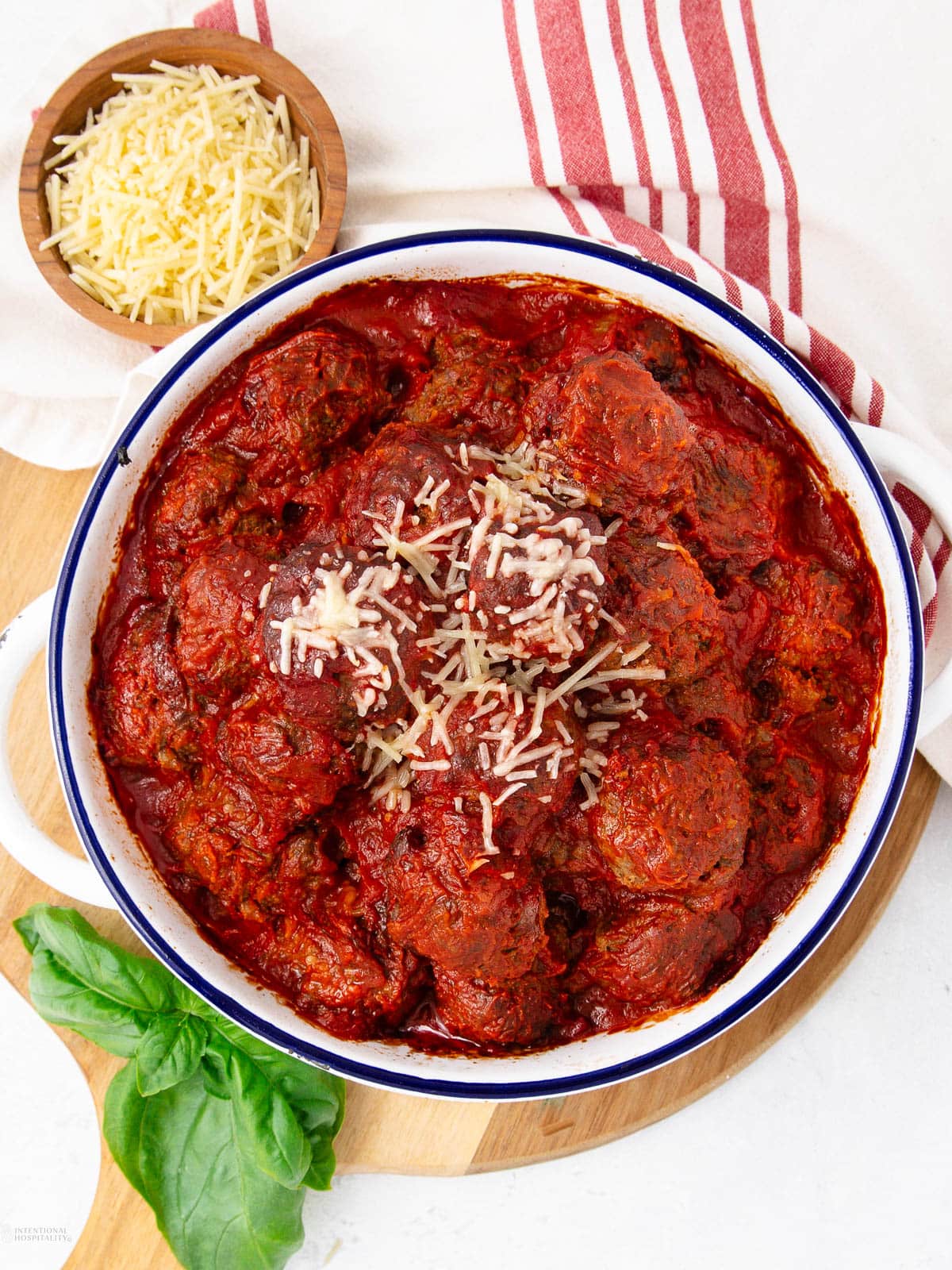 A round white dish filled with meatballs in marinara sauce, topped with shredded cheese. Nearby are a bowl of shredded cheese, a red and white striped towel, fresh basil leaves, and a wooden board.