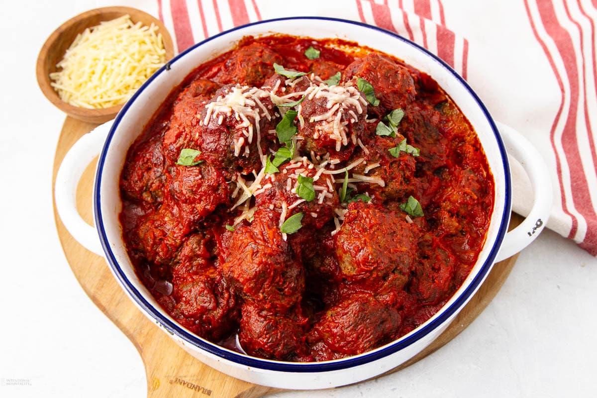 A bowl of meatballs covered in tomato sauce, garnished with shredded cheese and fresh basil, sits on a wooden board. A small bowl of extra cheese and a red-striped napkin are beside it.