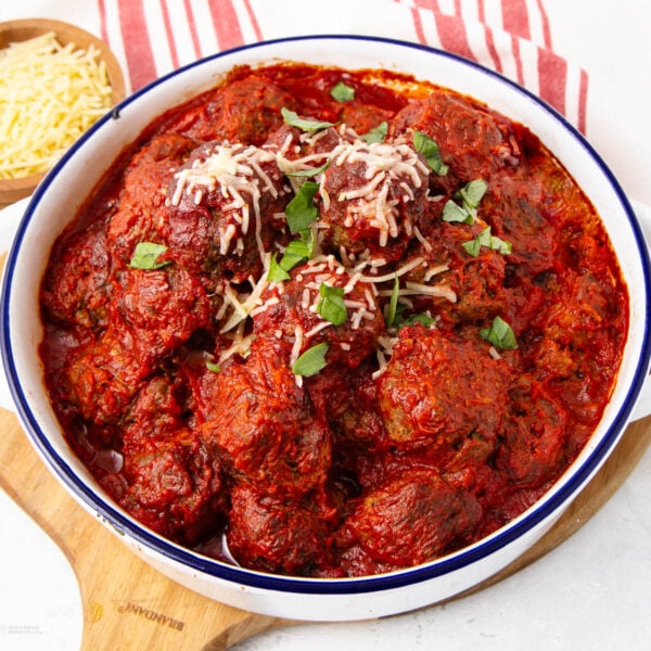 A white serving dish filled with meatballs covered in tomato sauce, topped with shredded cheese and fresh basil, sits on a wooden board next to a small bowl of grated cheese and a red-striped cloth.