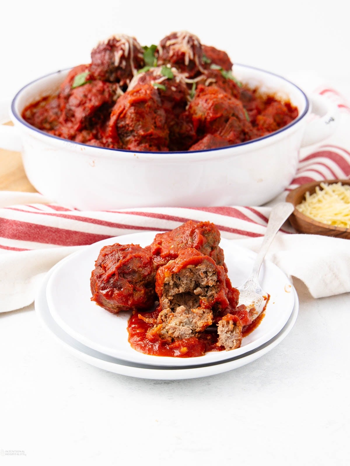 A plate with two large meatballs topped with tomato sauce, one cut open with a fork. In the background, a dish filled with more saucy meatballs and a striped red and white towel.
