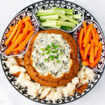 A platter with a bread bowl filled with creamy spinach dip, topped with chopped green onions, surrounded by chunks of bread, carrot sticks, and celery sticks arranged in separate sections.