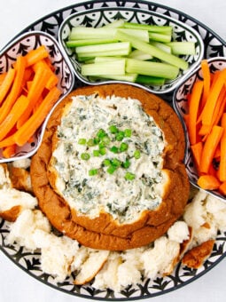 A platter with a bread bowl filled with creamy spinach dip, topped with chopped green onions, surrounded by chunks of bread, carrot sticks, and celery sticks arranged in separate sections.