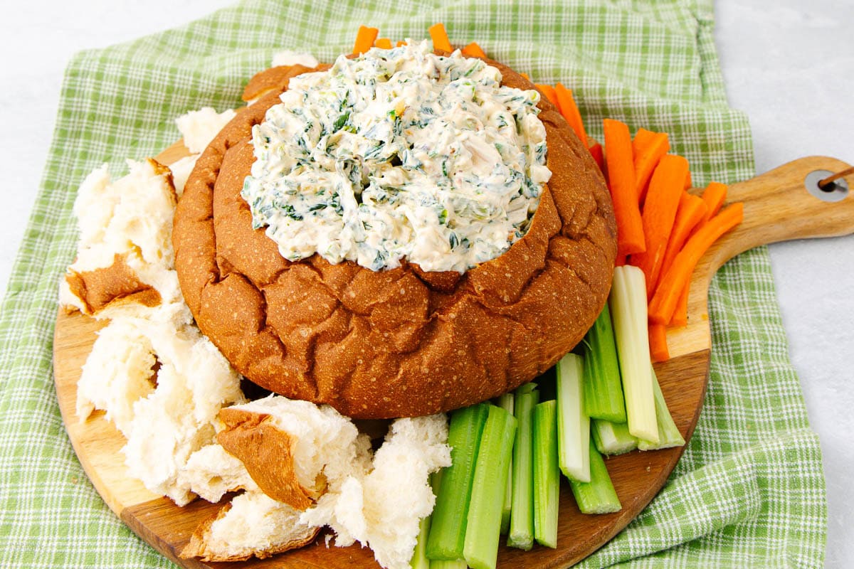 A round bread loaf hollowed out and filled with creamy spinach dip, served on a wooden board with celery sticks, carrot sticks, and chunks of bread, all placed on a green checkered cloth.