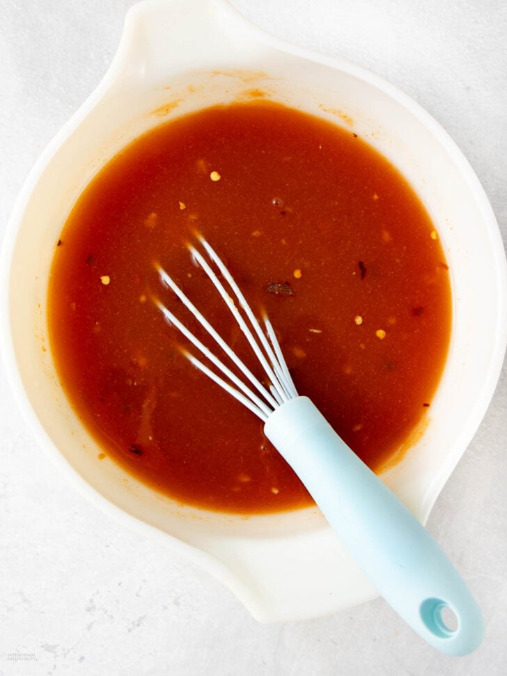 A white mixing bowl filled with a reddish-brown sauce and visible chili flakes, with a blue-handled whisk resting inside.
