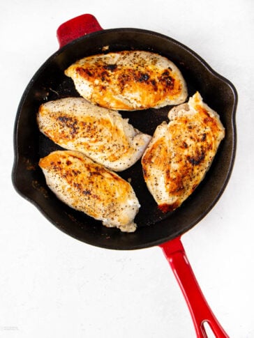 Four seasoned, golden-brown chicken breasts are cooking in a black cast iron skillet with a red handle, set against a white background.