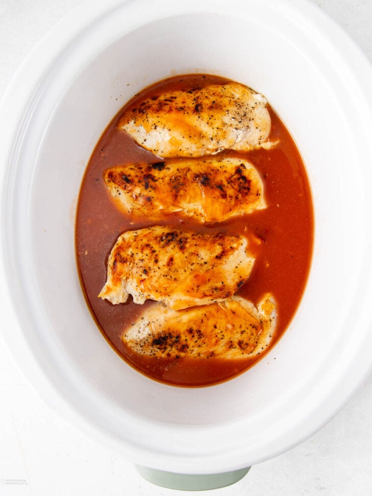 Four cooked chicken breasts in a white slow cooker, sitting in a reddish-brown sauce. The chicken is seasoned and the sauce surrounds the meat evenly.