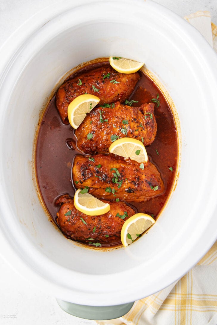 Four cooked chicken breasts in a slow cooker, covered in a rich, dark sauce, topped with chopped herbs and garnished with lemon slices. A yellow and white cloth is partially visible next to the slow cooker.