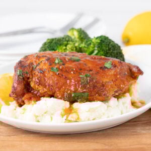 A plate with glazed chicken breast on mashed potatoes, garnished with herbs, served with broccoli. A lemon wedge and cutlery are in the background.
