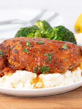 A plate with glazed chicken breast on mashed potatoes, garnished with herbs, served with broccoli. A lemon wedge and cutlery are in the background.