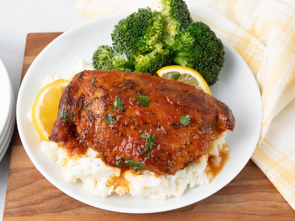 A plate with mashed potatoes topped with a seasoned baked chicken breast, garnished with parsley and lemon slices, served with steamed broccoli. A yellow-striped napkin is beside the plate.
