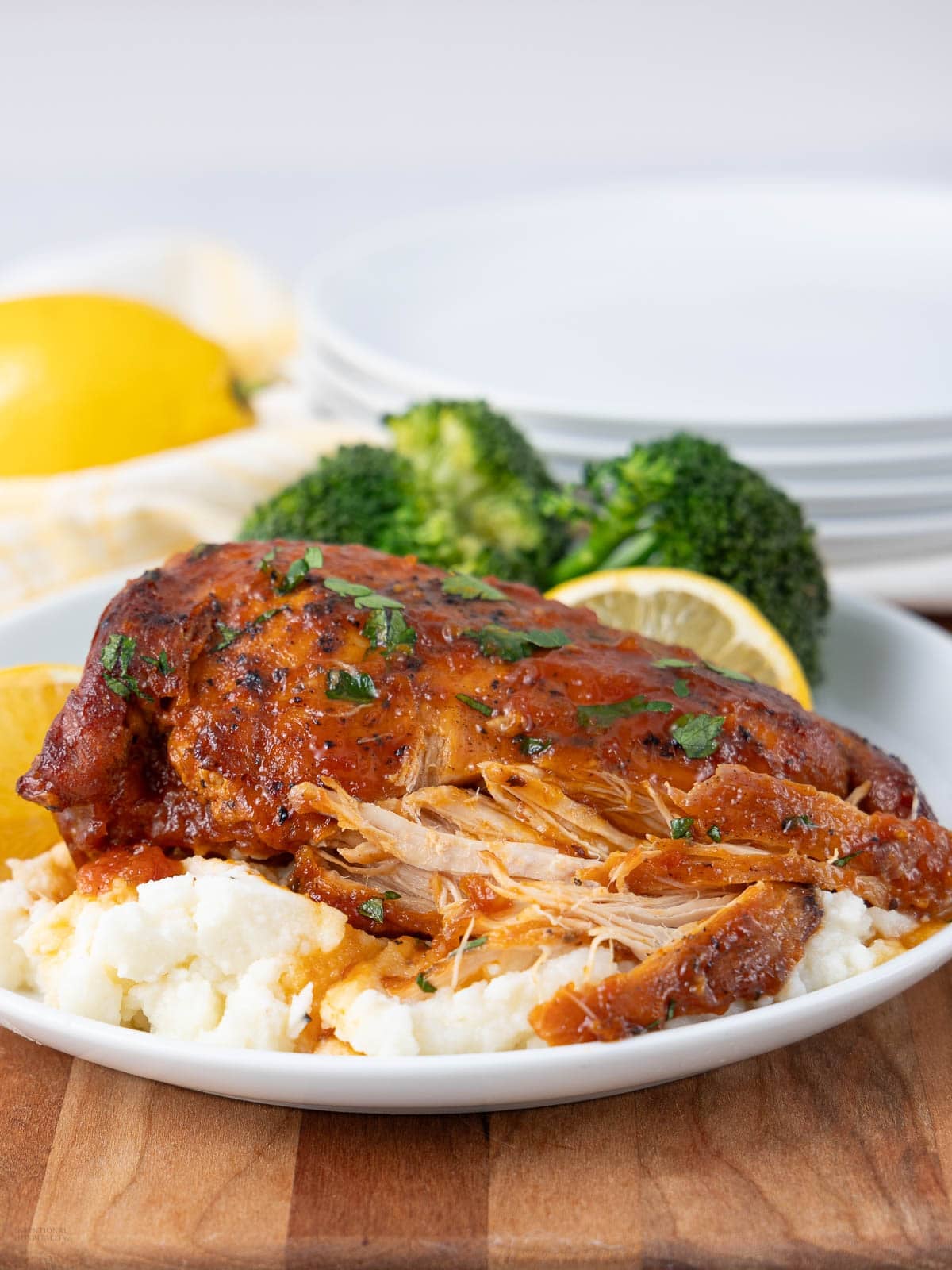 A plate of shredded, glazed chicken served over mashed potatoes, garnished with parsley and lemon slices, with steamed broccoli and stacked white plates in the background.