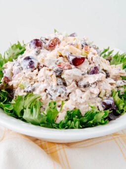 A plate of chicken salad with grapes, nuts, and creamy dressing is served on a bed of green lettuce leaves, placed on a white plate over a yellow-checked cloth.