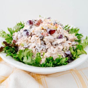 A plate of chicken salad with grapes, nuts, and creamy dressing is served on a bed of green lettuce leaves, placed on a white plate over a yellow-checked cloth.