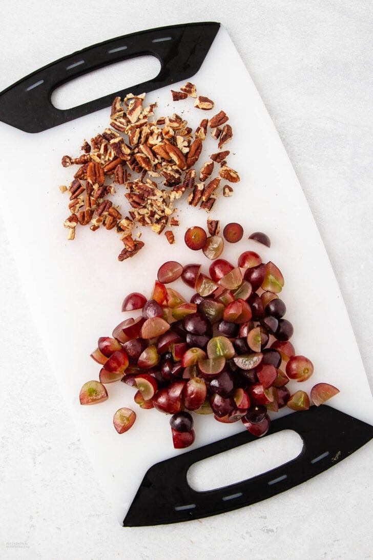 A white cutting board with black handles holds chopped pecans at the top and halved red grapes at the bottom, both on a light gray countertop.