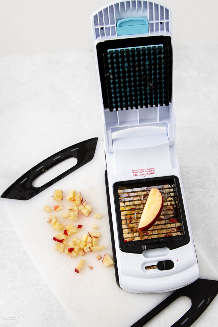 A white and black vegetable chopper with a sliced apple piece placed on its grid, sitting on a cutting board scattered with small, diced apple pieces.