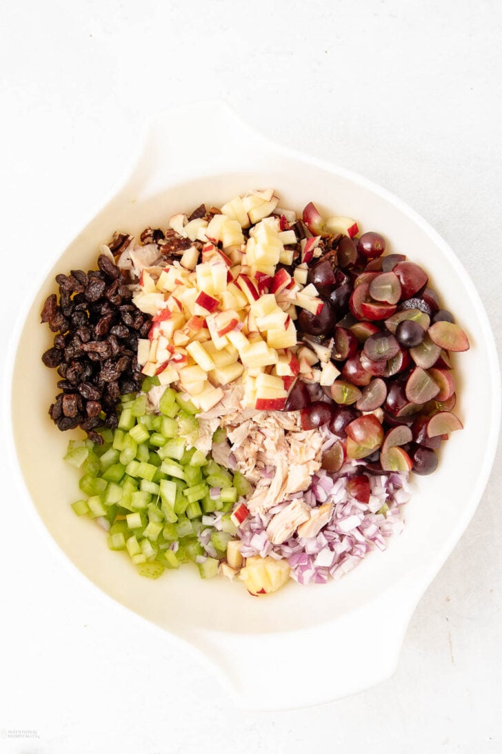 A white bowl filled with neatly arranged ingredients: chopped apples, halved grapes, diced celery, shredded chicken, diced red onion, and raisins, all on a white background.