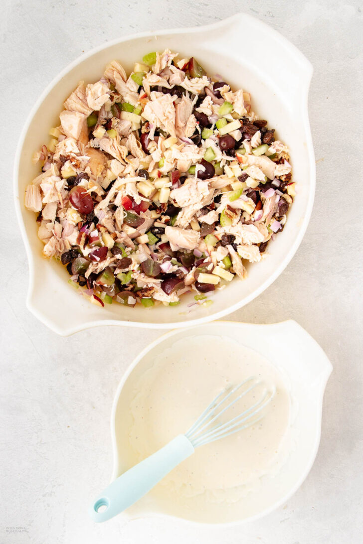 A bowl of chopped chicken salad with grapes, celery, apple, and nuts sits above another bowl containing a creamy dressing being whisked with a blue whisk on a light surface.