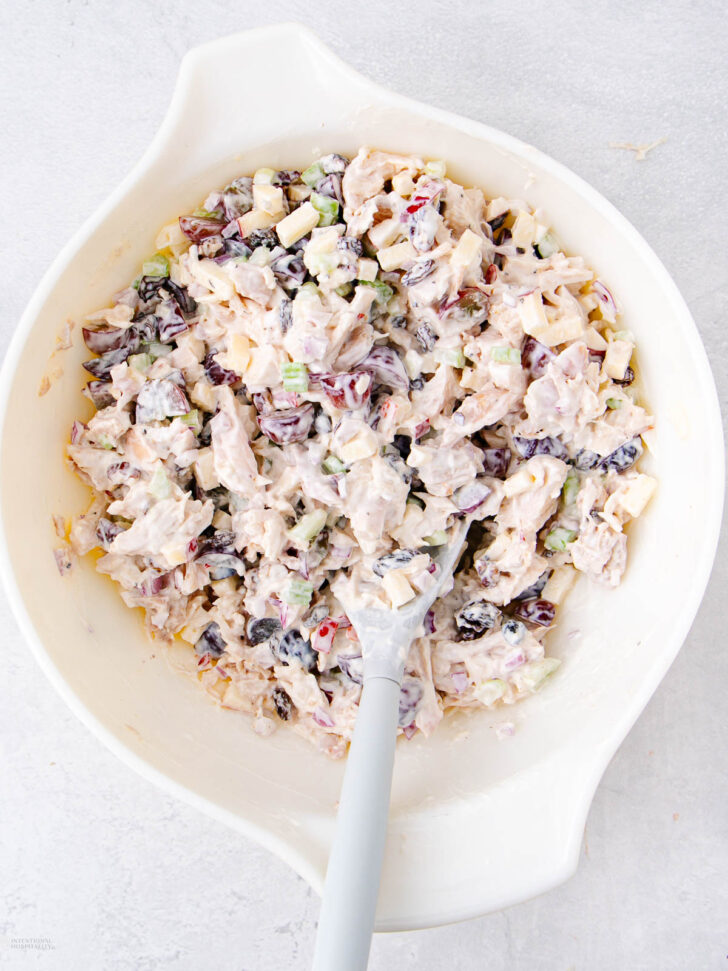 A white mixing bowl filled with creamy chicken salad containing chunks of chicken, celery, apple, dried cranberries, and slivered almonds. A spatula is resting in the bowl.