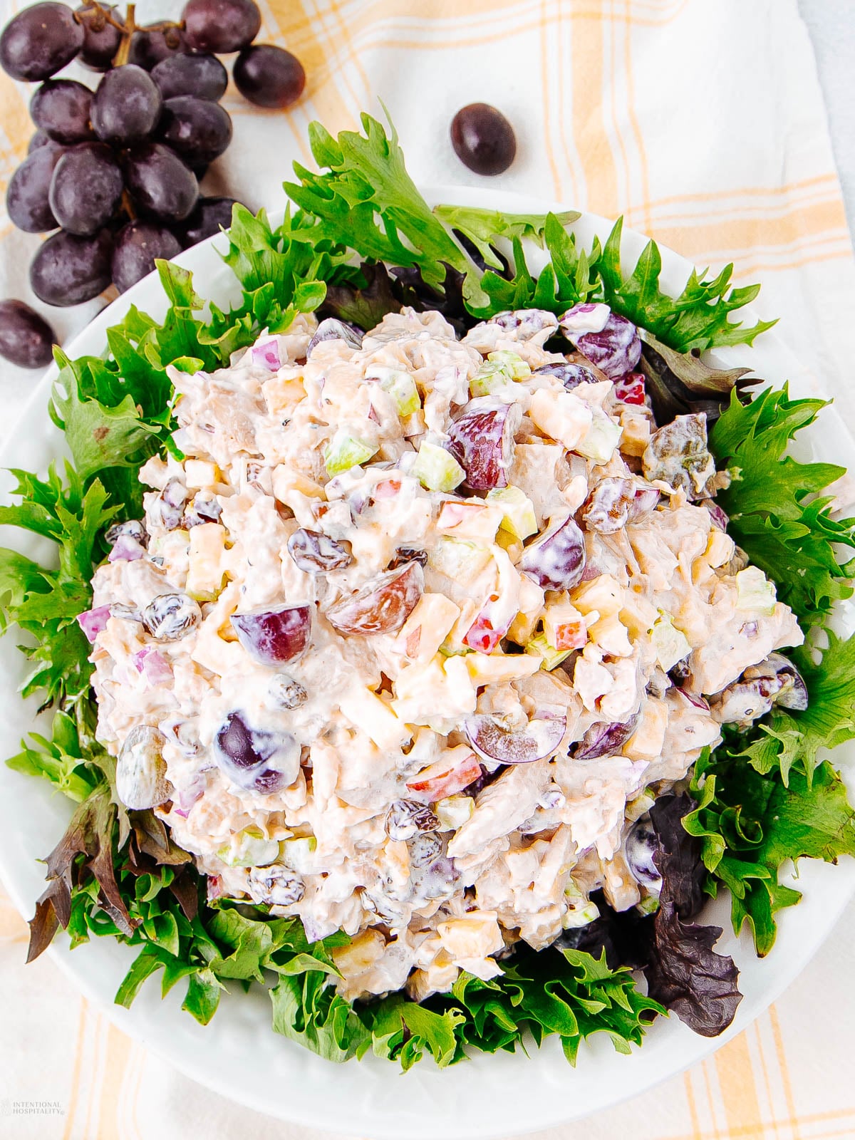 A bowl of creamy chicken salad with grapes, celery, and chopped nuts, served on a bed of leafy greens. A bunch of purple grapes and a yellow plaid napkin are in the background.