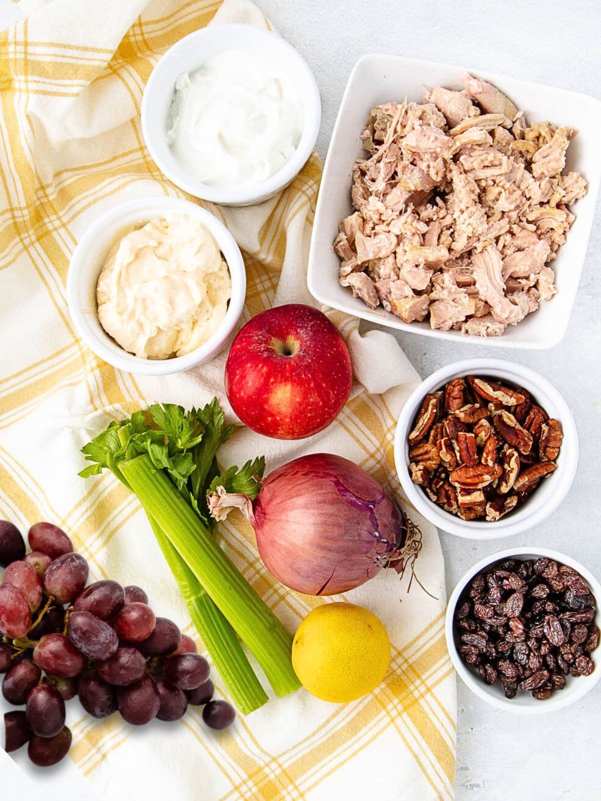 A flat lay of ingredients for waldorf chicken salad on a yellow plaid cloth, featuring bowls of yogurt, mayonnaise, shredded chicken, pecans, and raisins with a red apple, celery stalks, red onion, lemon, and red grapes.