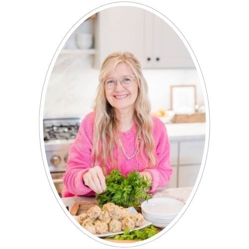 A smiling woman with long blonde hair and glasses, wearing a bright pink sweater, prepares fresh herbs and food in a modern kitchen.