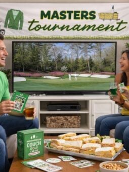 Four people sit on couches, smiling and holding drinks and snacks, watching the Masters golf tournament on TV. A banner reading "Masters Tournament" hangs above, with sandwiches and snacks on the table.