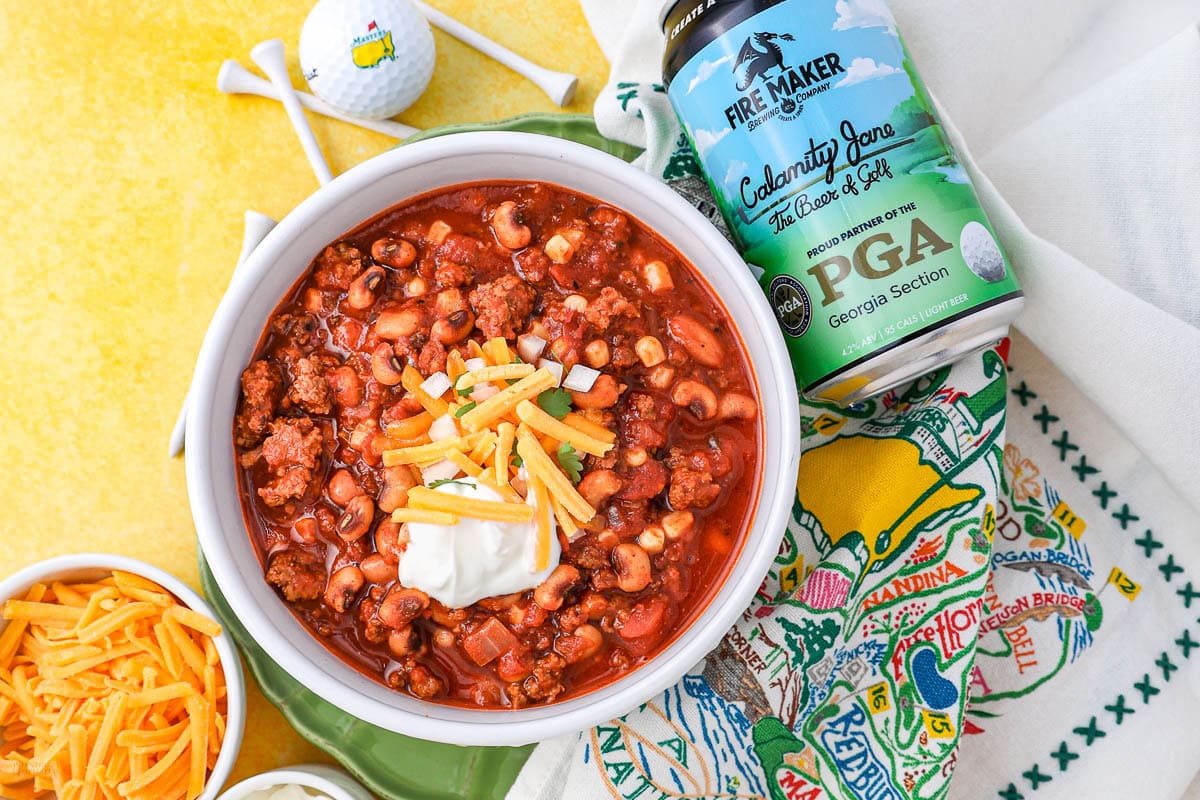 A bowl of chili with macaroni, ground meat, corn, and beans, topped with shredded cheese and sour cream, sits on a colorful golf-themed napkin next to a can of beer, shredded cheese, golf tees, and a golf ball.