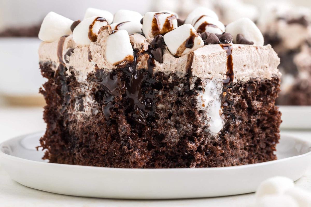 A close-up of a slice of chocolate poke cake topped with whipped cream, chocolate drizzle, mini marshmallows, and chocolate chips on a white plate.