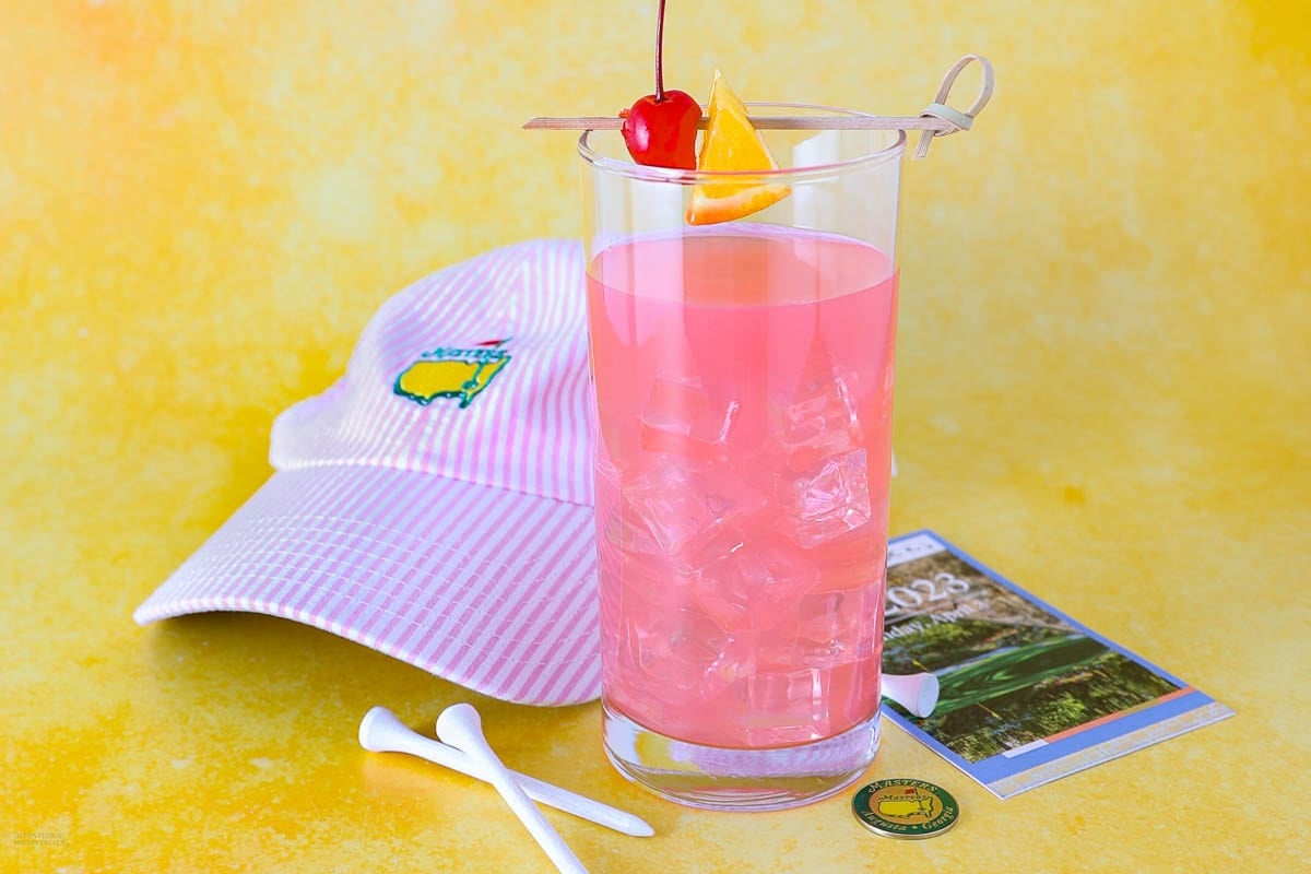 A tall glass of pink iced drink garnished with a cherry and orange slice, sits in front of a pink-striped hat with a green and yellow logo. A couple of golf tees and a ticket are nearby on a yellow background.
