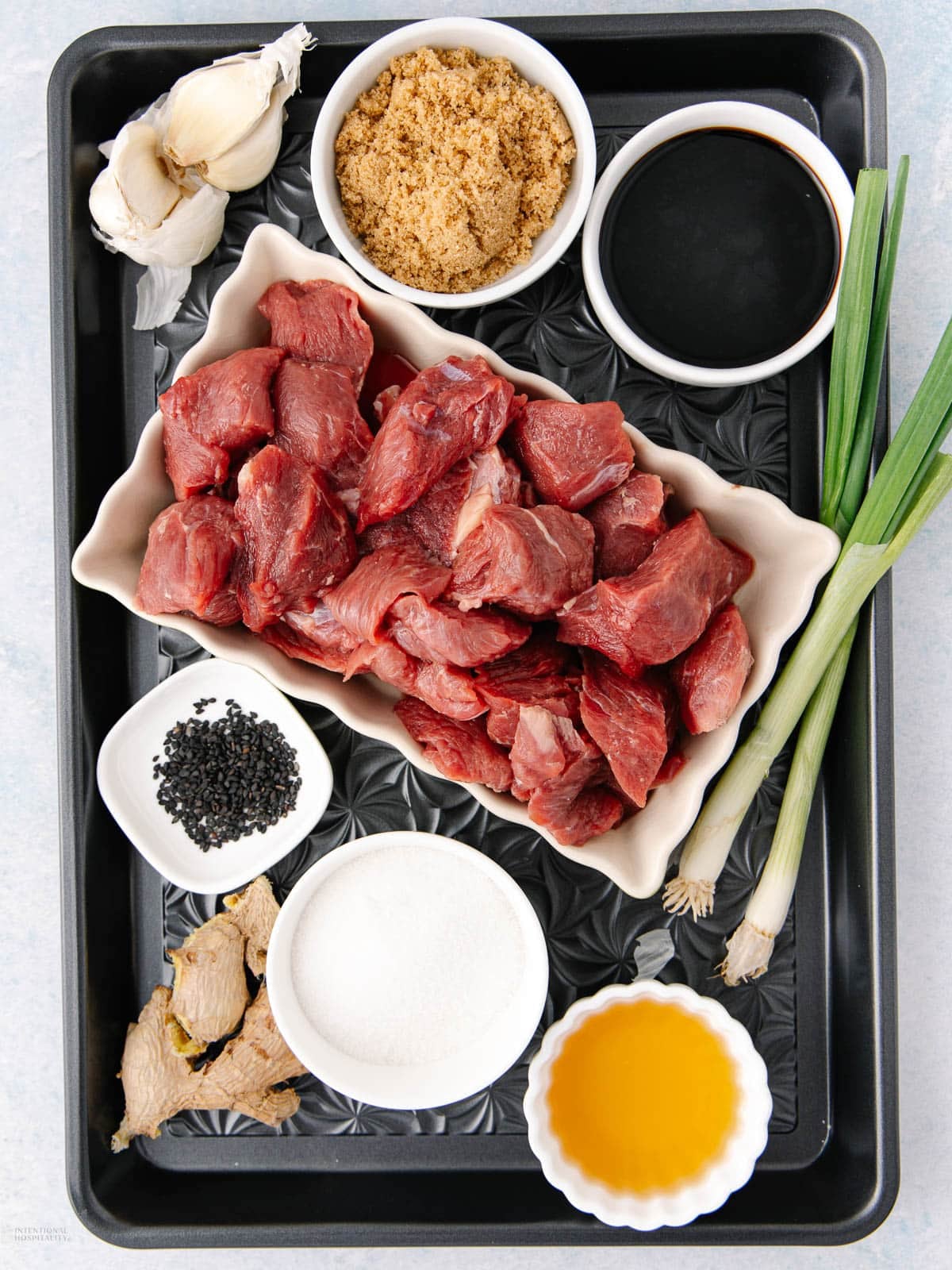 A tray with raw beef cubes, brown sugar, soy sauce, green onions, garlic cloves, ginger slices, black sesame seeds, white sugar, and a small bowl of yellow liquid, arranged in separate bowls and plates.