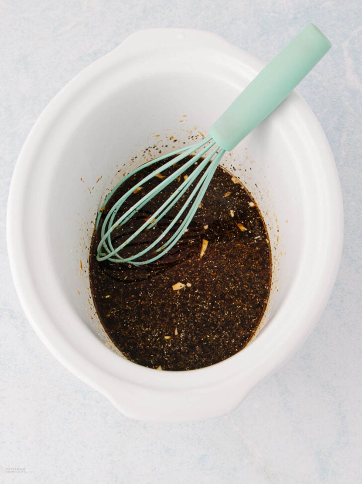 A white slow cooker containing a dark brown marinade with visible spices and garlic pieces, and a light blue whisk resting inside.