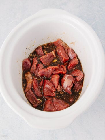 Chunks of raw beef marinating in a dark sauce inside a white slow cooker on a light-colored surface.
