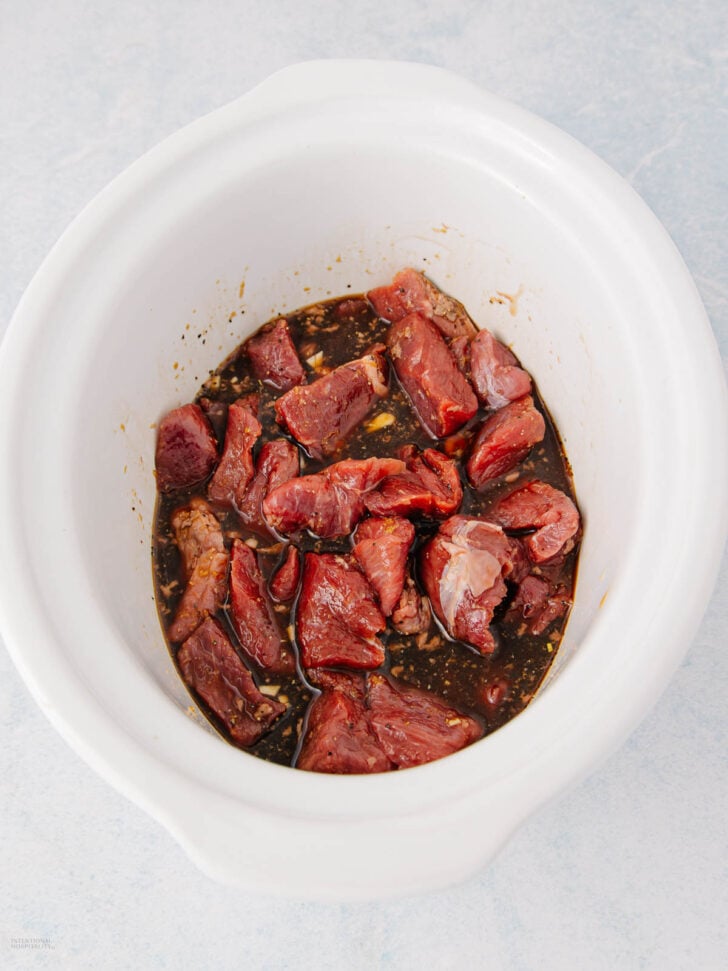Chunks of raw beef marinating in a dark sauce inside a white slow cooker on a light-colored surface.