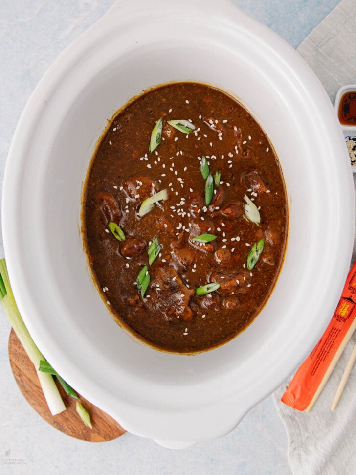 A white slow cooker filled with beef in a dark brown sauce, garnished with sliced green onions and sesame seeds. Nearby are chopsticks, a sauce dish, a napkin, and green onions on a wooden board.