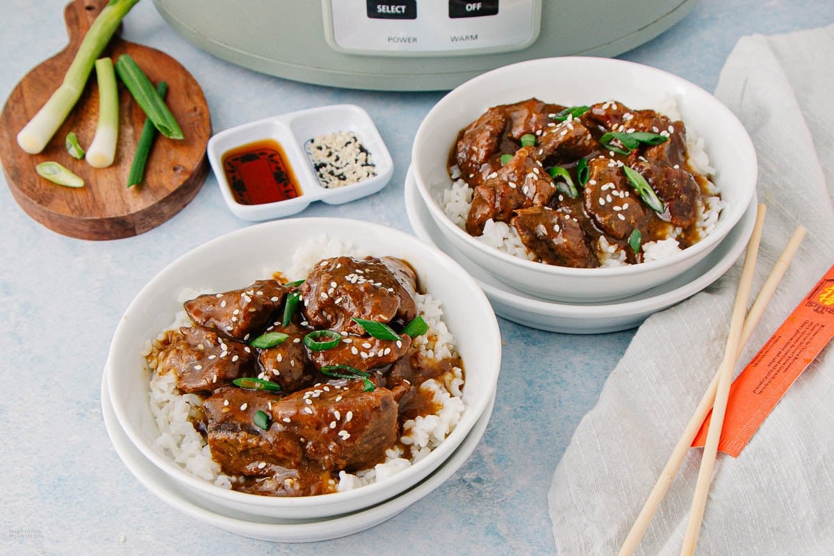 Two white bowls filled with rice and glazed beef topped with sesame seeds and sliced green onions. Nearby are chopsticks, a cloth napkin, sauces, and a small cutting board with sliced green onions.