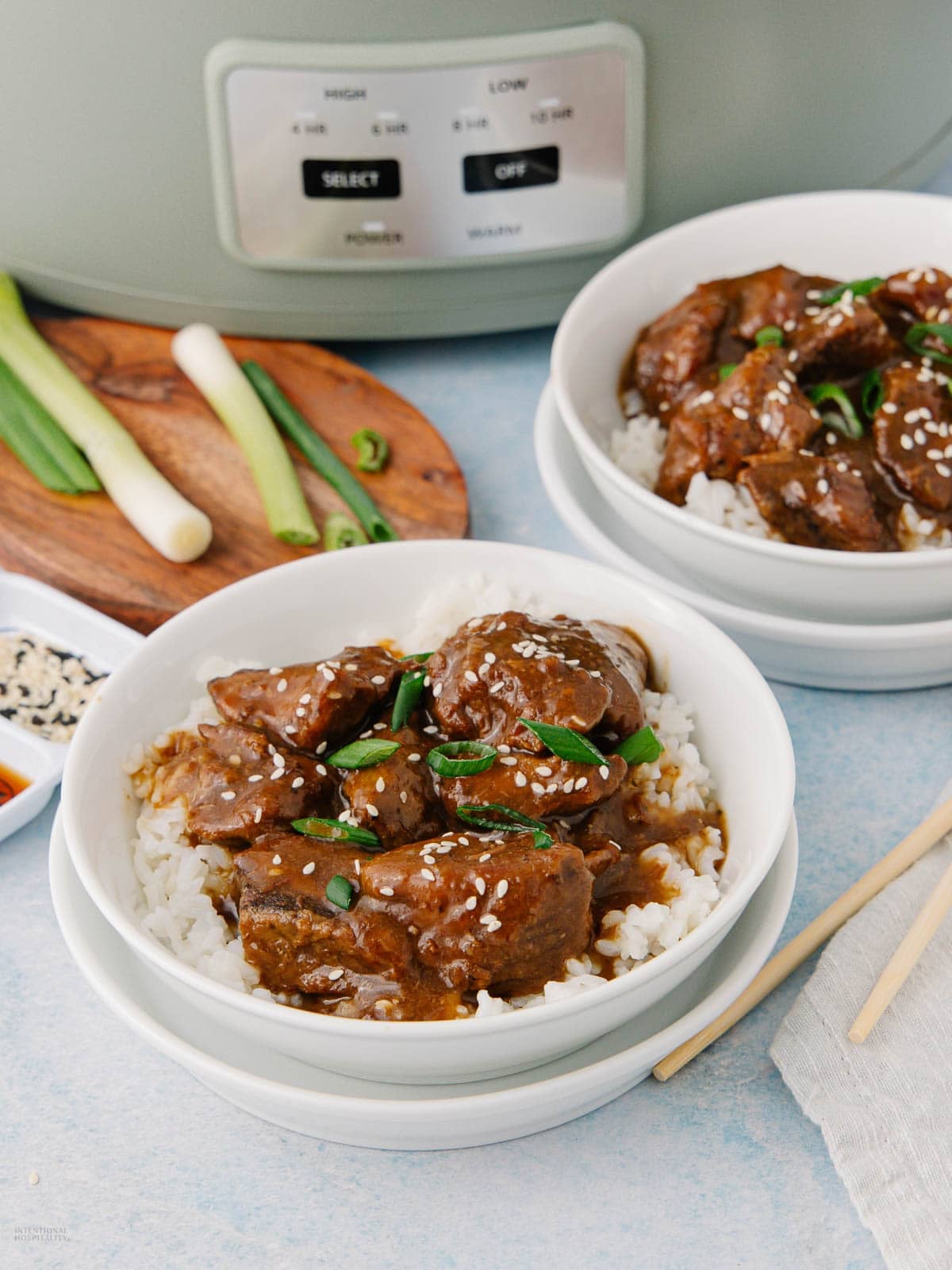 Two bowls of white rice topped with glazed beef, sliced green onions, and sesame seeds sit on a table near a slow cooker, with green onions and a small dish of sesame seeds nearby.
