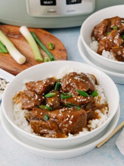 Two white bowls filled with rice and tender beef in a brown sauce, garnished with sesame seeds and sliced green onions. Green onions, sauce, and a slow cooker are in the background. Chopsticks rest beside the bowls.
