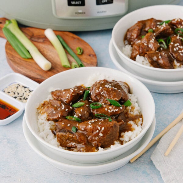 Two white bowls filled with rice and tender beef in a brown sauce, garnished with sesame seeds and sliced green onions. Green onions, sauce, and a slow cooker are in the background. Chopsticks rest beside the bowls.