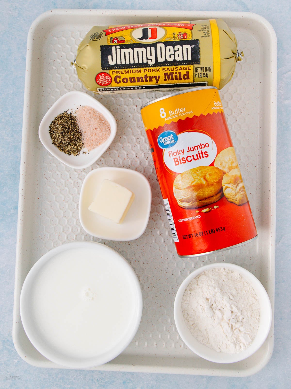 Tray with a roll of Jimmy Dean country mild pork sausage, a can of Great Value flaky jumbo biscuits, a dish of butter, two small bowls with salt and pepper, a bowl of flour, and a bowl of milk.