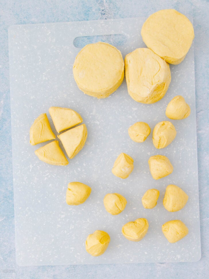 Round pieces of biscuit dough, some whole and some cut into smaller portions, are arranged on a white cutting board against a light blue surface.