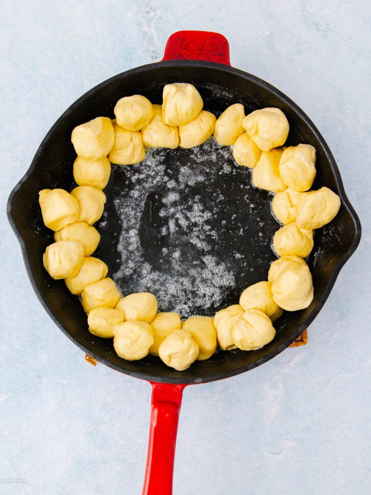 A black cast iron skillet with a red handle contains small dough balls arranged in a ring around the edge, with the center empty. The skillet sits on a light blue surface.