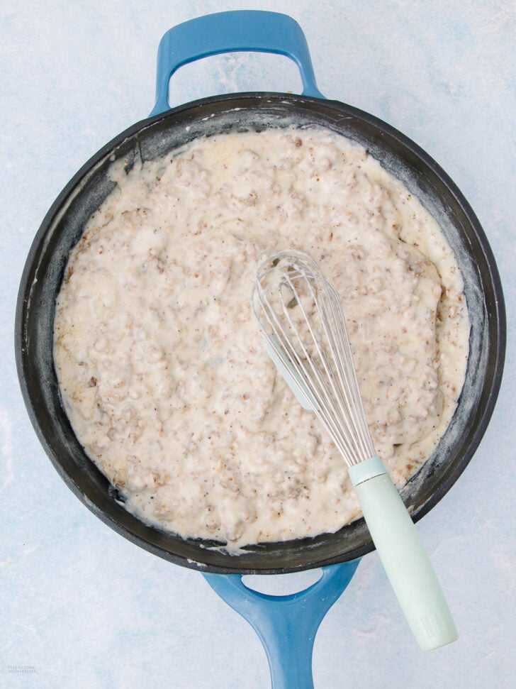 A blue-handled skillet filled with creamy sausage gravy, with a whisk resting inside the mixture, sits on a light blue surface.