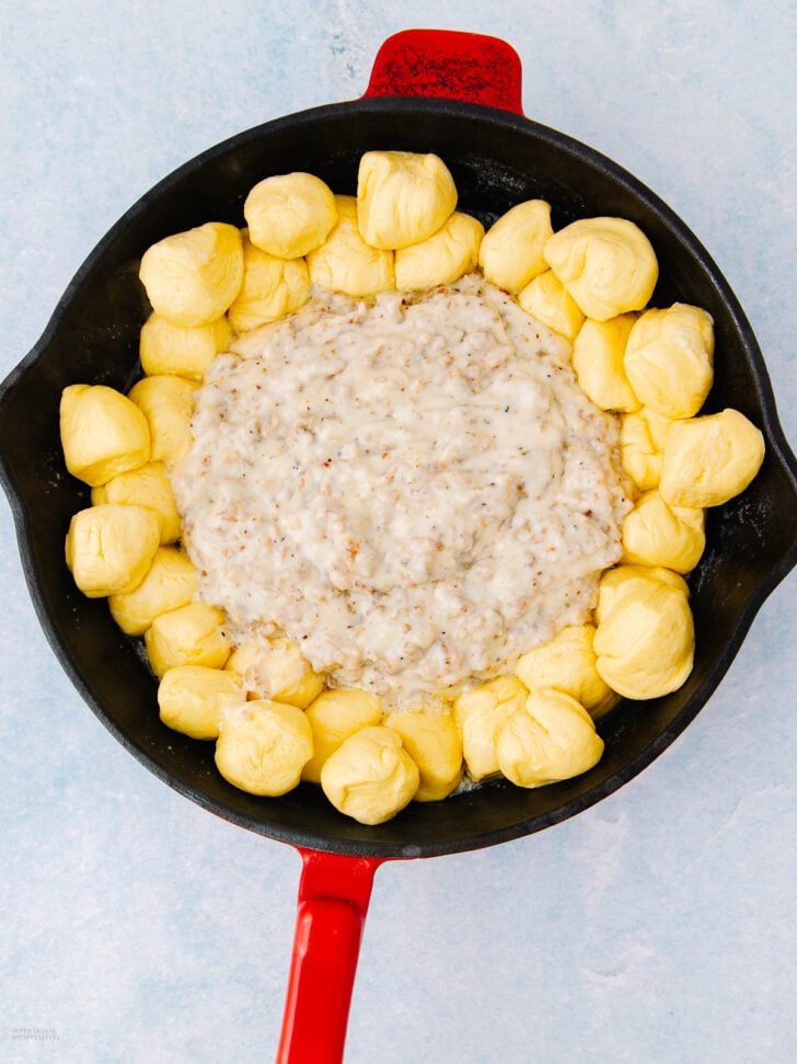 A cast iron skillet with a red handle contains biscuit dough balls arranged in a circle around the edge, with creamy sausage gravy in the center. The background is a light blue surface.