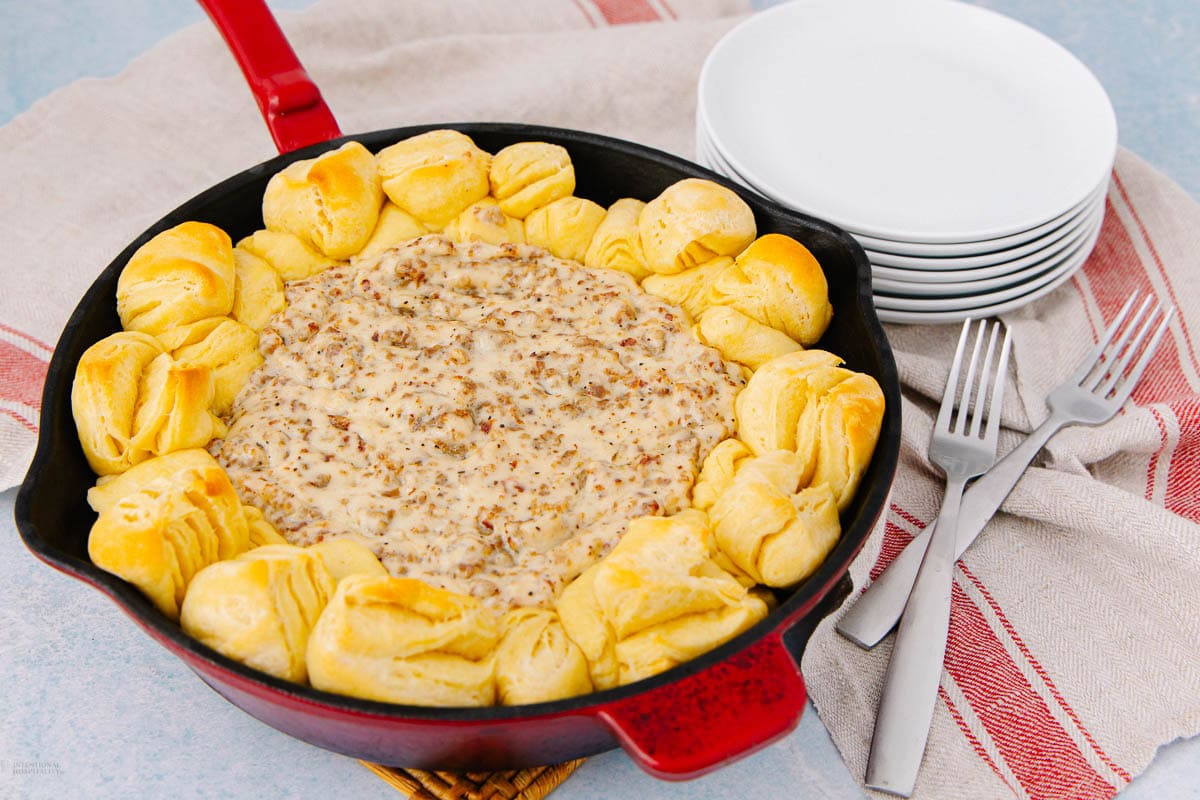 A skillet filled with creamy sausage gravy in the center, surrounded by golden baked biscuits. Nearby are a stack of white plates and forks on a red-striped cloth.
