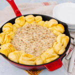 A red skillet filled with creamy sausage dip in the center, surrounded by golden baked biscuit pieces, sits on a woven mat next to a stack of white plates, forks, and a striped towel.