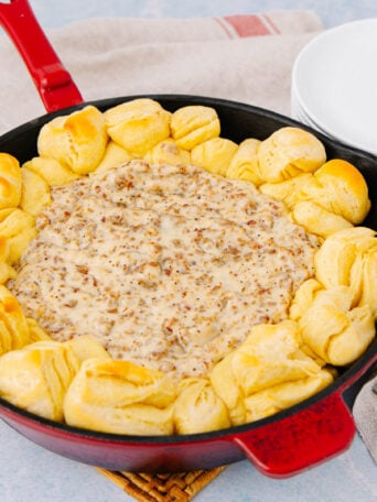 A red skillet filled with creamy sausage dip in the center, surrounded by golden baked biscuit pieces, sits on a woven mat next to a stack of white plates, forks, and a striped towel.
