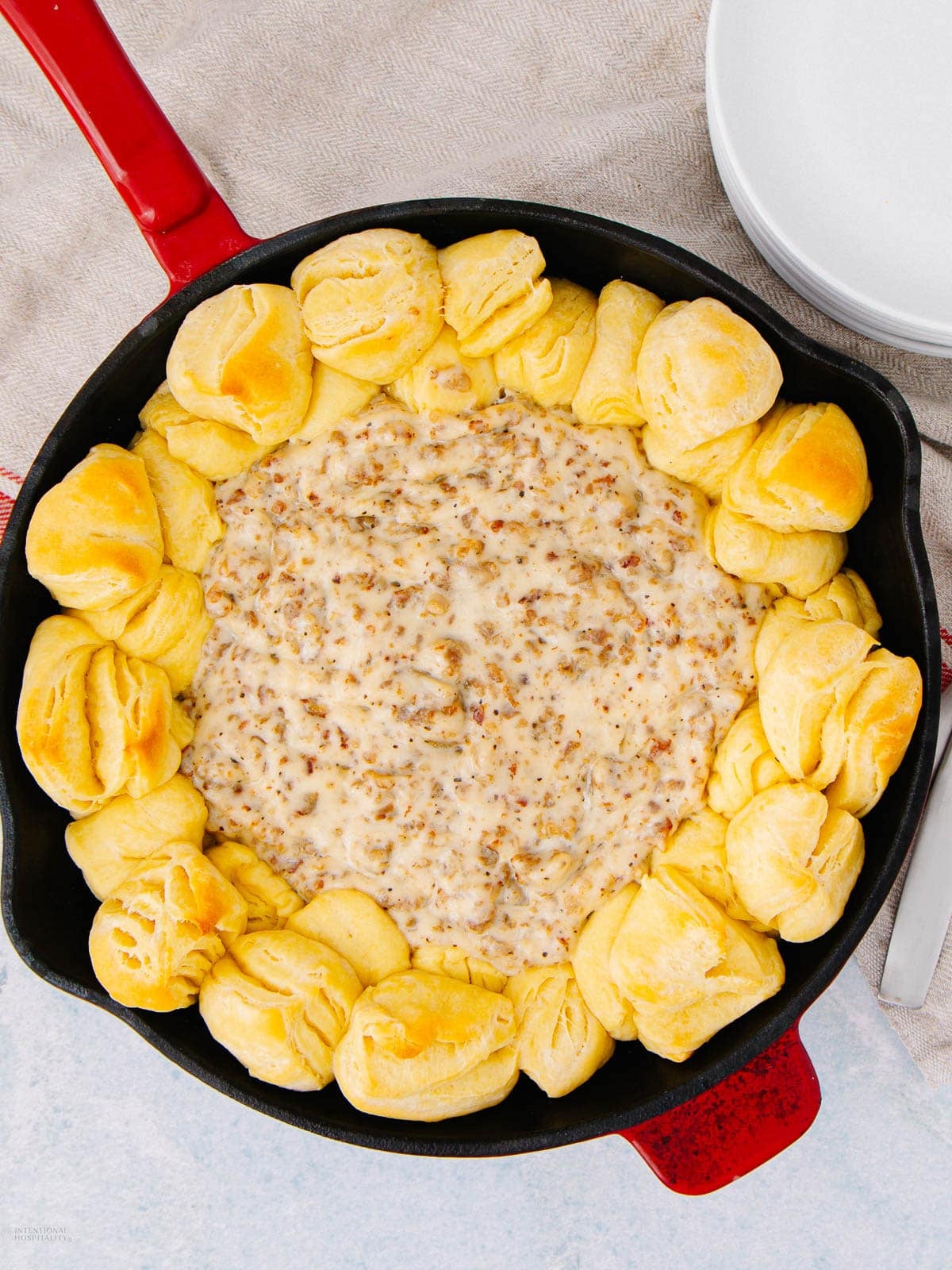 A red skillet filled with creamy sausage gravy in the center, surrounded by golden, baked biscuit pieces around the edge. The skillet sits on a light cloth next to a stack of white plates.